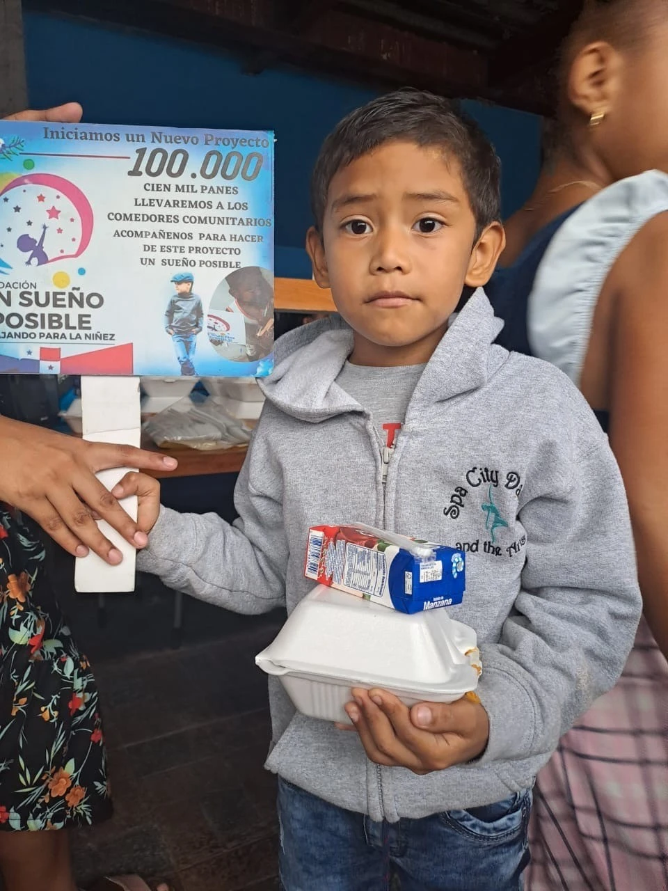 Niño con alimentos junto al cartel del Proyecto de los 100.000 Panes